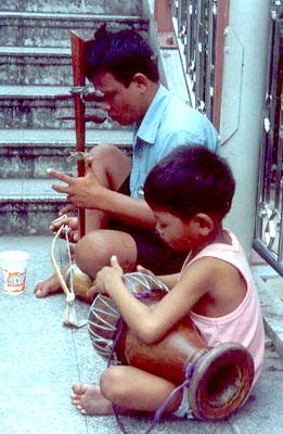 A boy beats on a drum while his father plays a Thai saw.